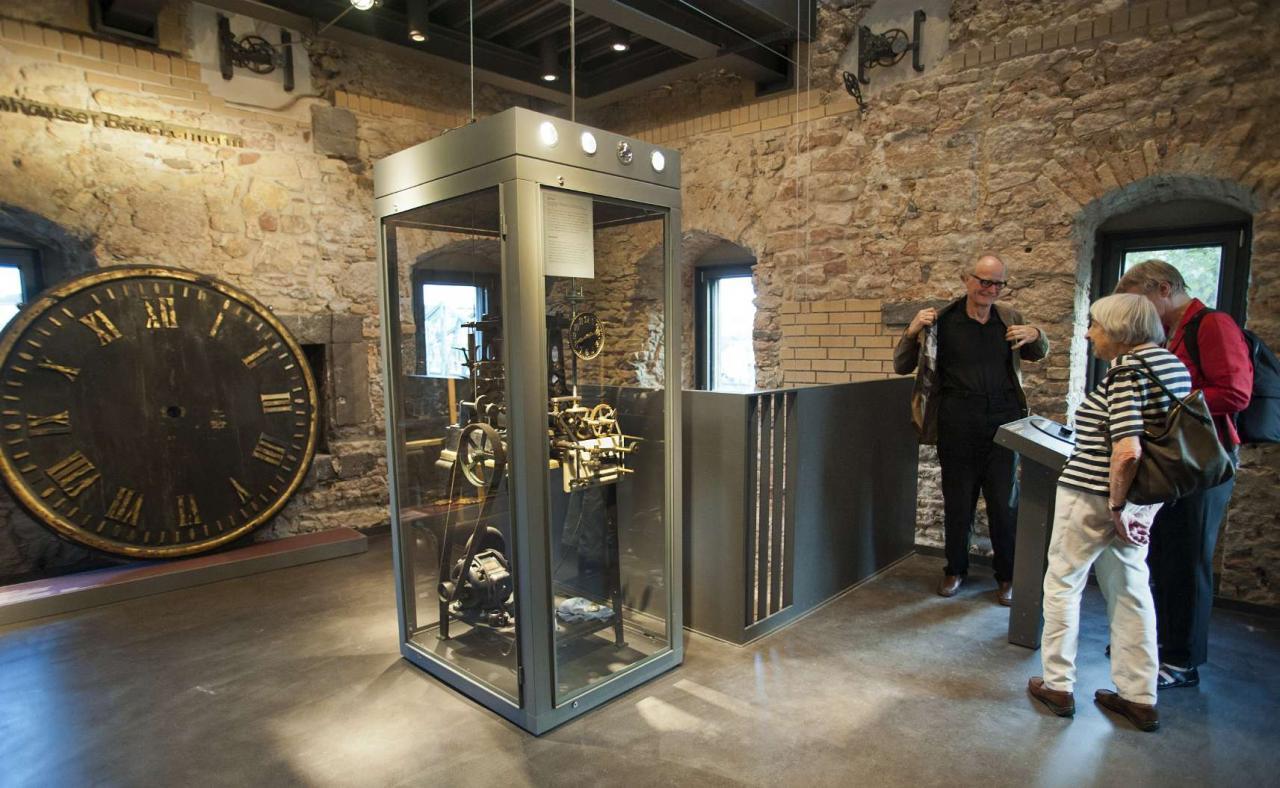 Das Bild zeigt einen Blick in den Rententurm, im Vordergrund steht ein Uhrwerk in einem Glaskasten, an der gegenüberliegenden Wand lehnt eine große analoge Uhr. 